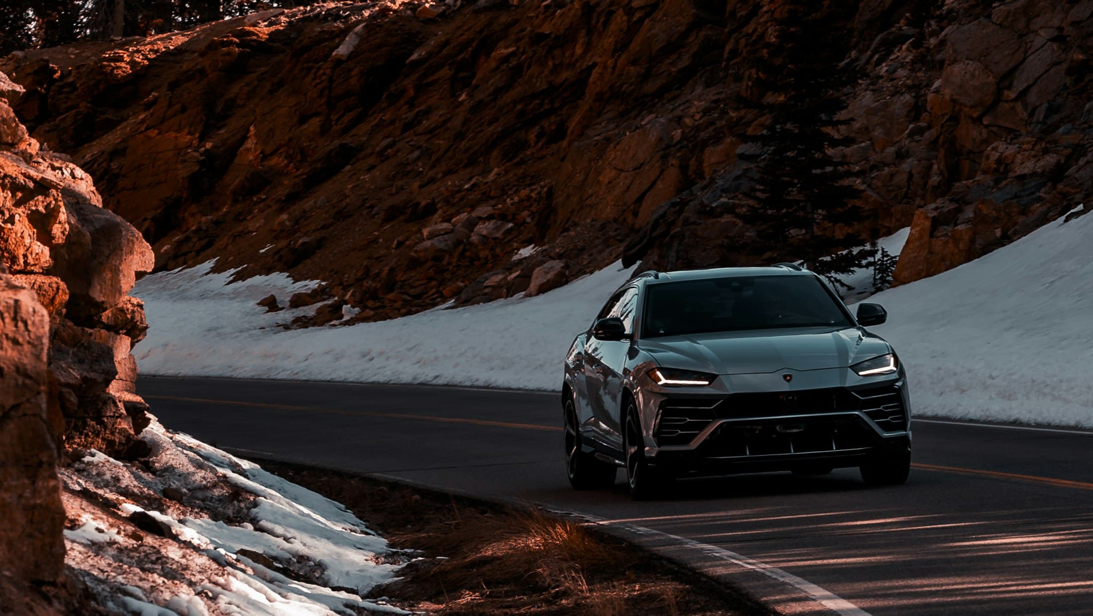 A grey SUV on a snow-lined mountain road below a rock face.
